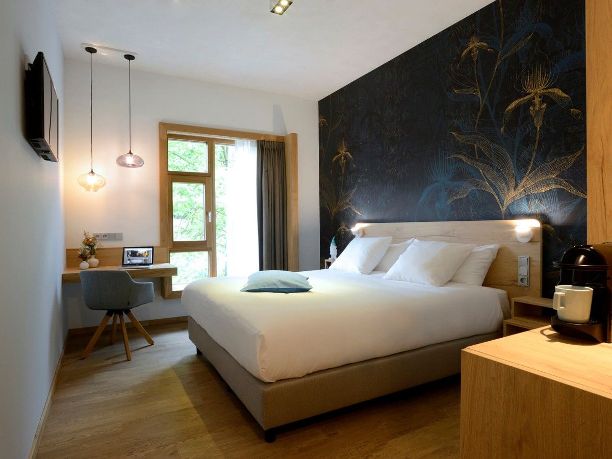 A modern hotel room with a large bed, a desk with a laptop, a coffee maker, and a wall with a floral design.