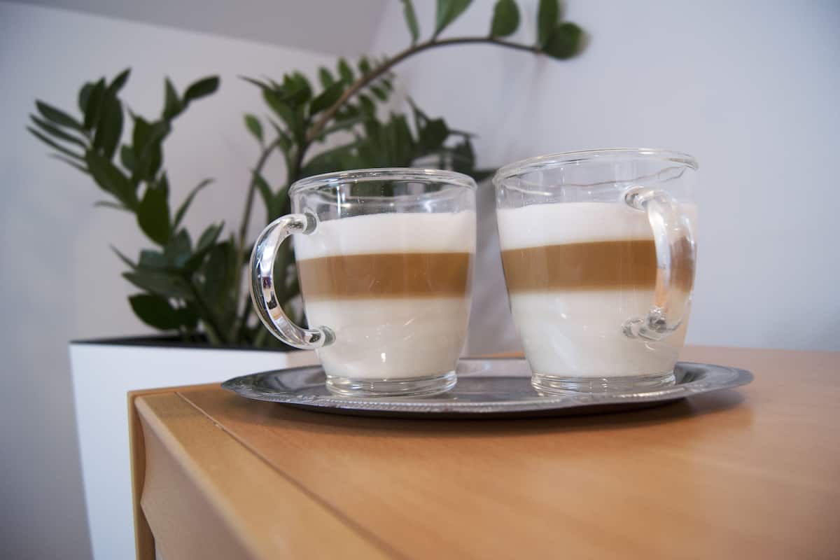 Two clear glass mugs with layered drinks on a coaster, placed on a wooden table.