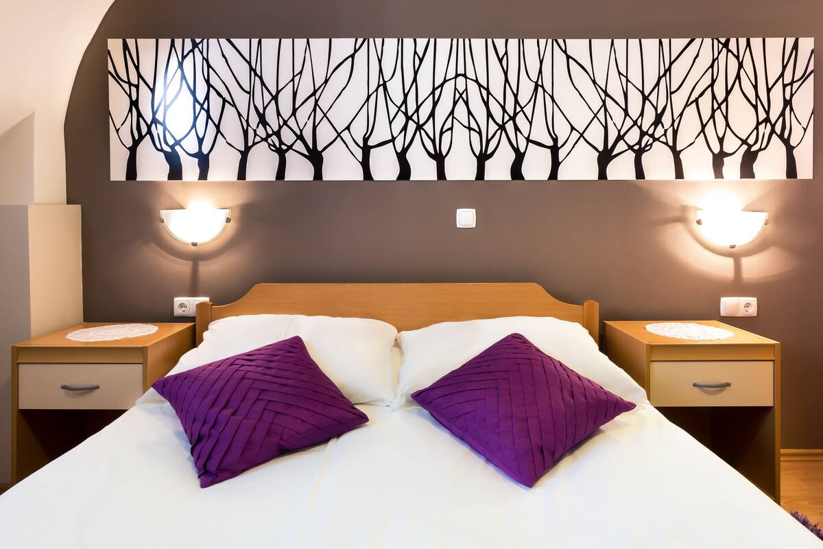A neatly made bed with two purple pillows, a wall-mounted artwork of bare trees, and bedside lamps on either side.