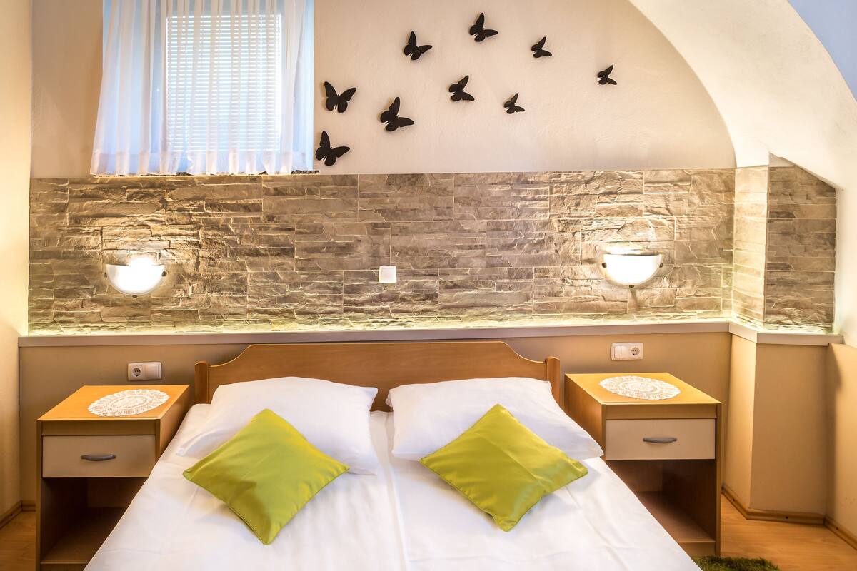 A bedroom with a stone wall, two bedside tables with lamps, a bed with green pillows, and a butterfly wall decoration.