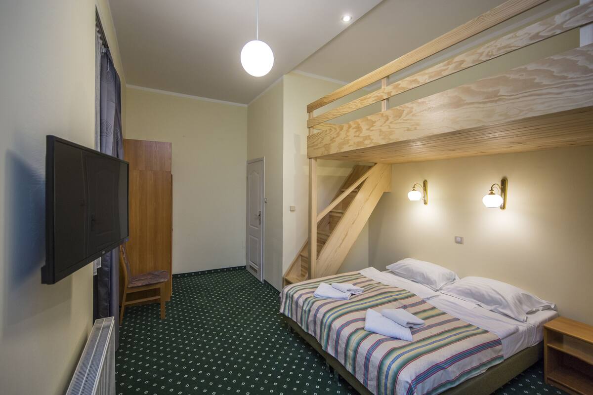 A room with a bunk bed, a flat-screen TV mounted on the wall, a wooden chair, a small desk, and a staircase.