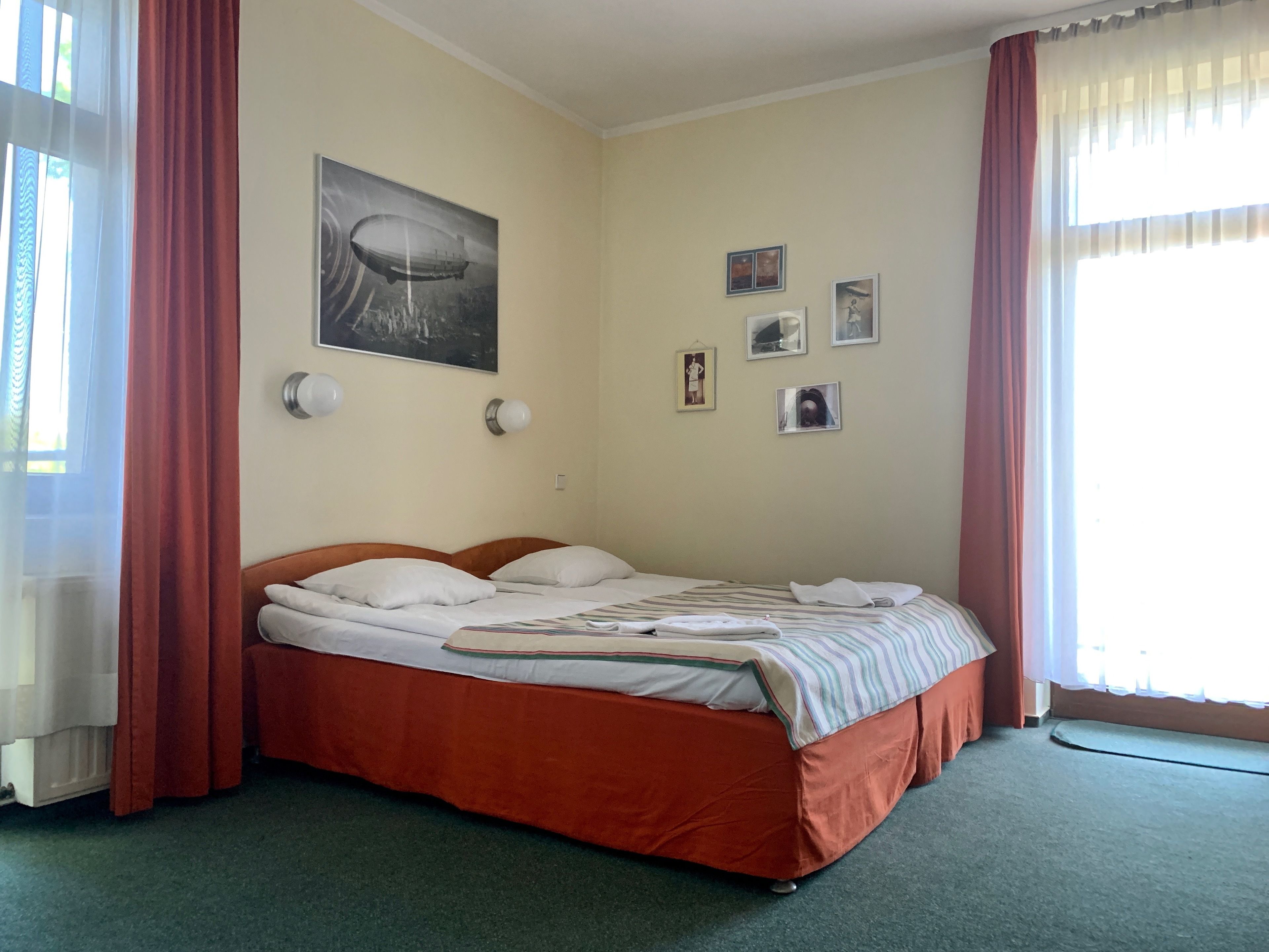 Uma cama bem arrumada com um edredom listrado, um quadro fixado na parede com o retrato de um dirigível e algumas pequenas molduras.