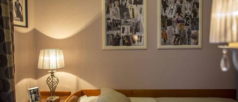 A hotel room with a neatly made bed, a bedside lamp, a framed collage on the wall, and a nightstand with a photo frame.