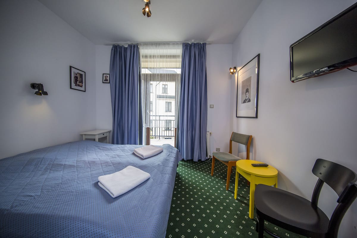 A hotel room with a bed, a chair, a small table, a TV, and a window with curtains.