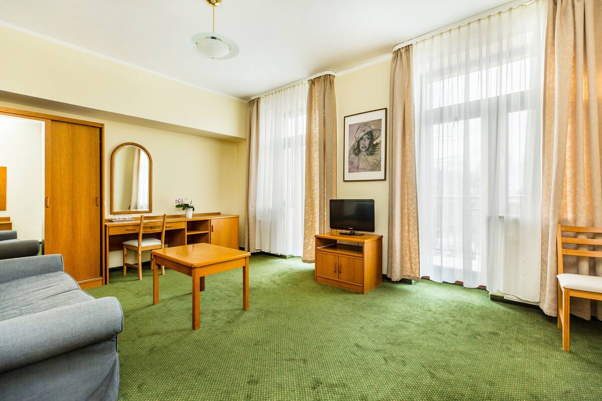 A hotel room with a green carpet, a wooden desk, a television, a sofa, and a large window with curtains.