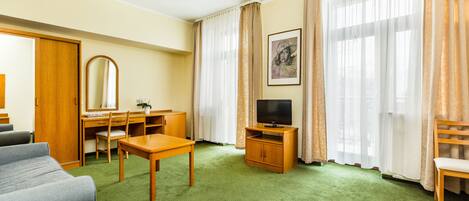 Quarto de hotel com carpete verde, escrivaninha de madeira, televisão, sofá e uma grande janela com cortinas.