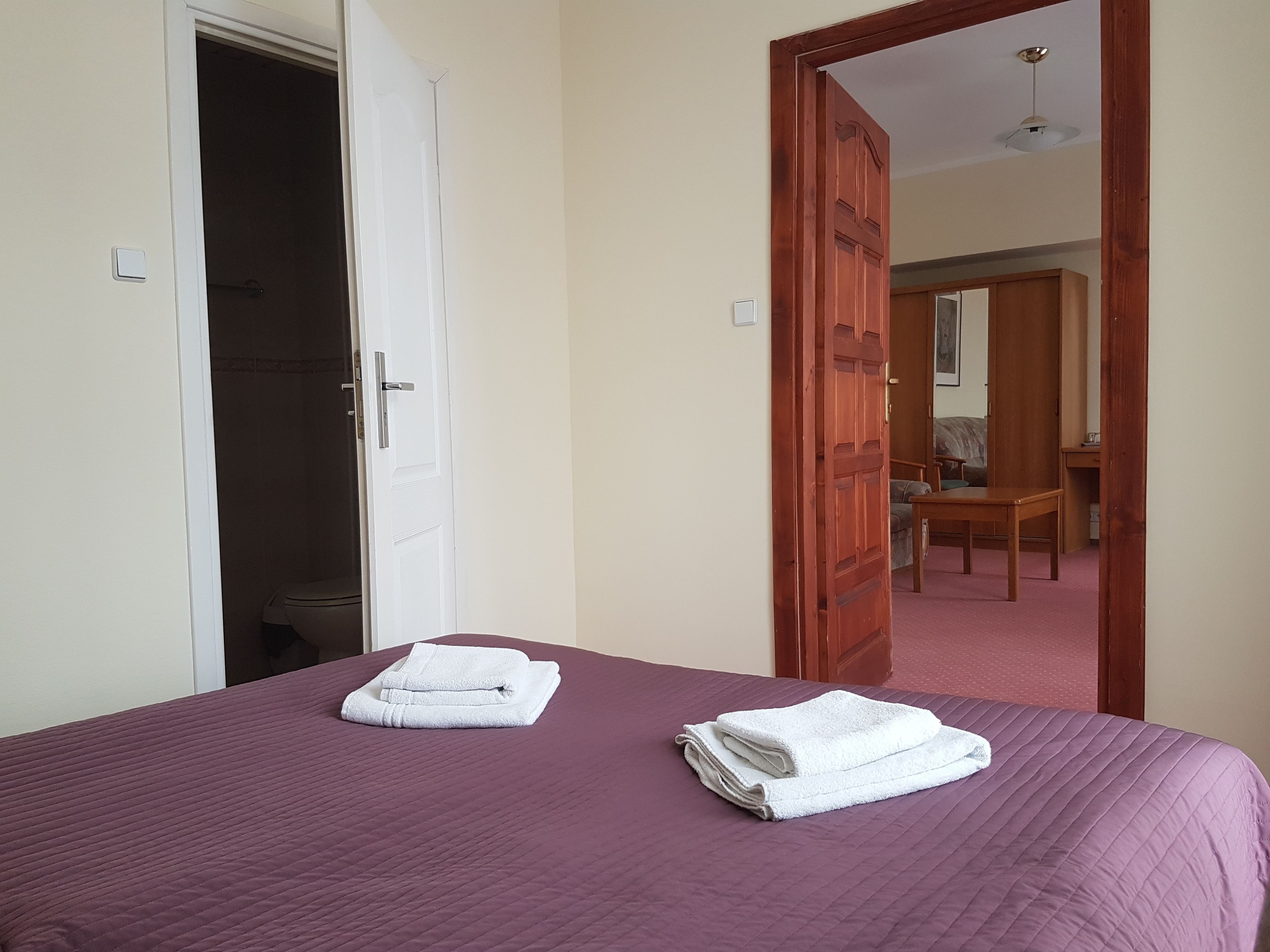 A hotel room with a bed, two white towels, a wooden door, and a view of a dining area.