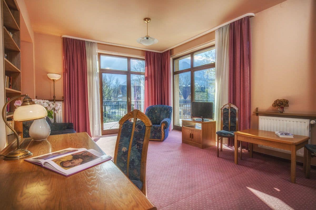 A cozy room with a wooden desk, a chair, a sofa, and a television. There are large windows with curtains, a radiator, and a bookshelf.