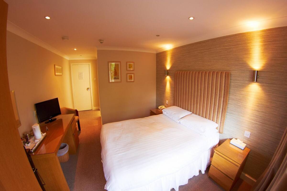A hotel room with a bed, bedside tables, a desk with a television, and wall-mounted lighting.