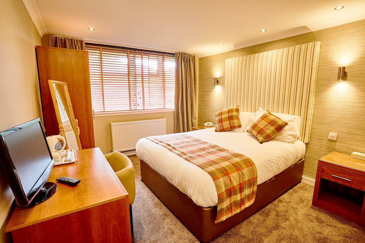 A hotel room with a large bed, a desk with a computer and a telephone, and a window with blinds.