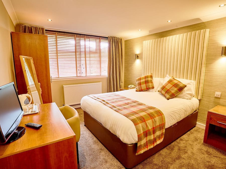 A hotel room with a large bed, a desk with a computer and a telephone, and a window with blinds.