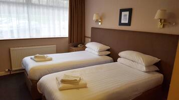 A hotel room with two beds, a radiator, a window with curtains, a bedside table with a lamp, and a framed picture on the wall.