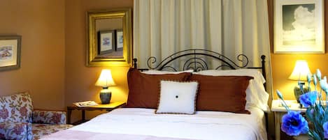 Habitación de hotel con una cama, una mesita de noche con lámpara, una silla, un cuadro enmarcado y un jarrón con flores.