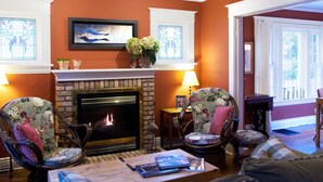 A living room with a fireplace, two floral armchairs, a coffee table with books, and a view of the outdoors through large windows. - Simcoe Suites on the Henley B&B (St. Catharines)