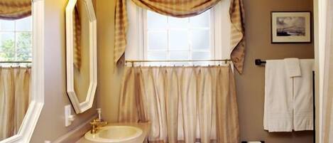 Un baño con dos lavamanos, un espejo, una ventana con cortinas, un inodoro y un perchero para toallas.