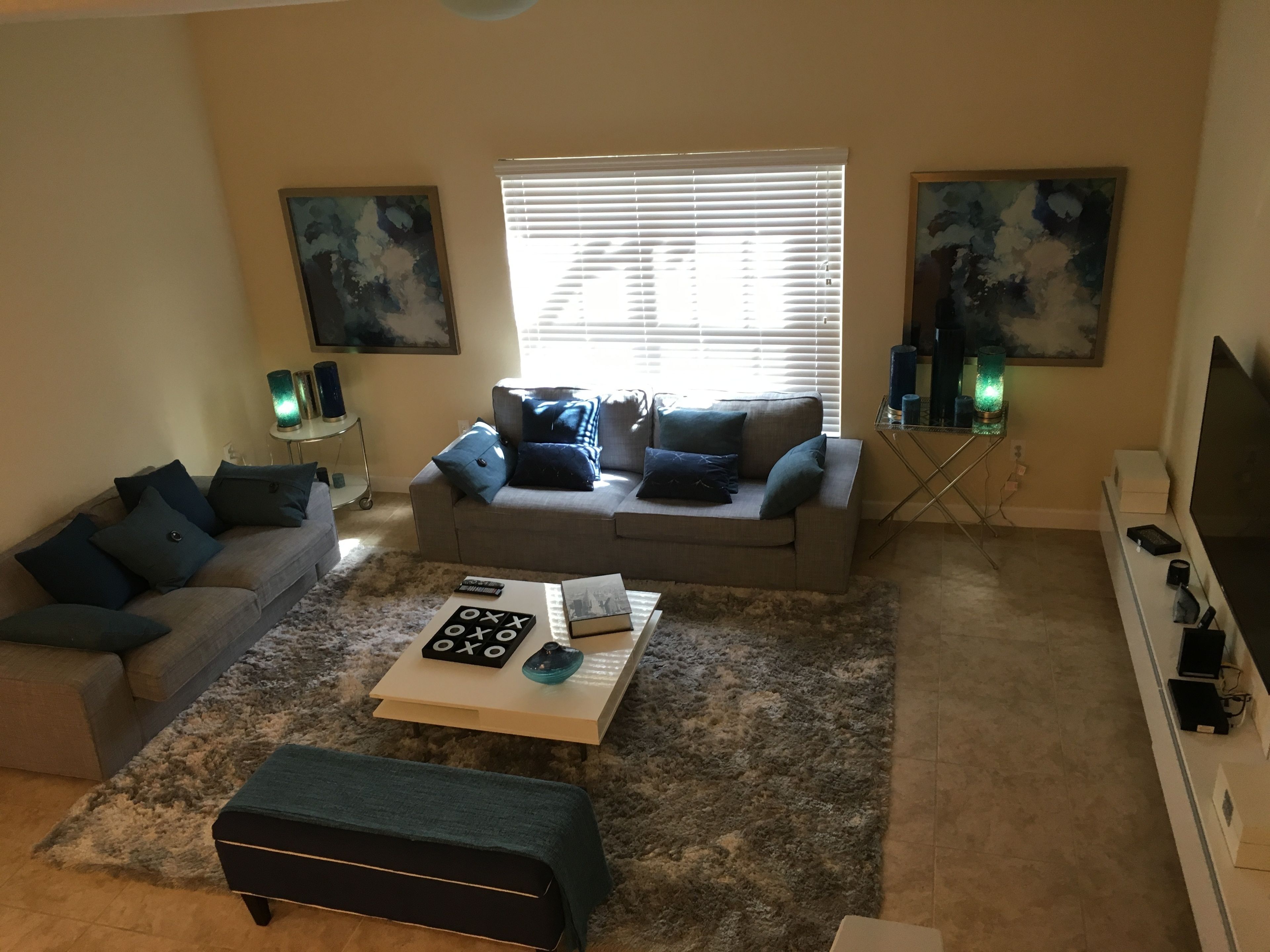 A living room with a grey sofa, a coffee table, and a TV stand. There are two paintings on the walls and a window with blinds.