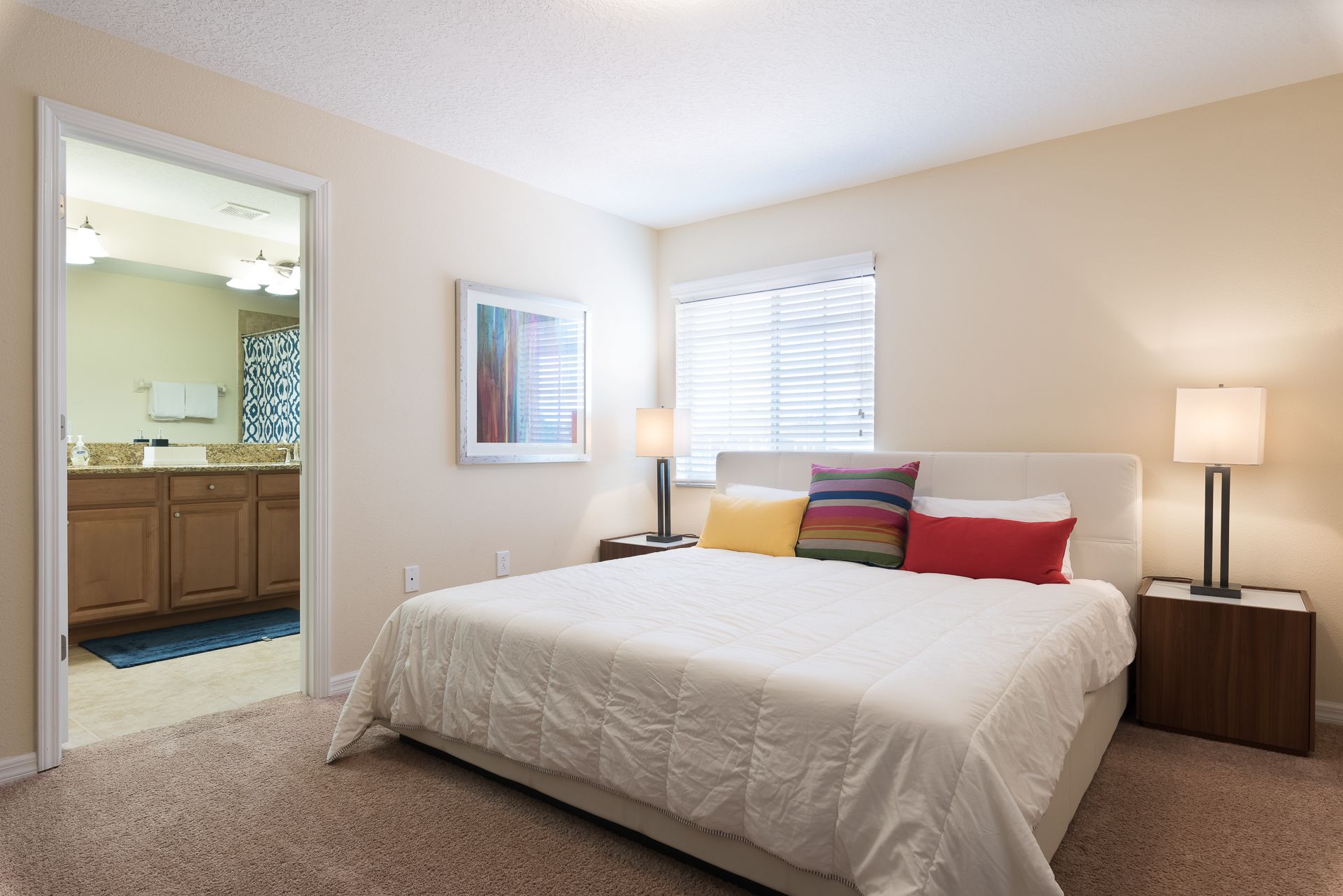 A bedroom with a large bed, two bedside tables with lamps, a window with blinds, a bathroom visible through an open door, and a framed picture on the wall.