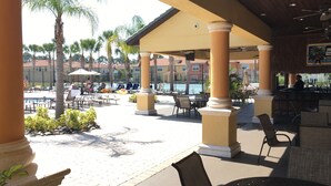 An outdoor pool area with lounge chairs and umbrellas, surrounded by palm trees and a bar area.