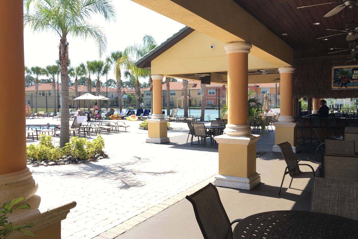 An outdoor pool area with lounge chairs and umbrellas, surrounded by palm trees and a bar area.