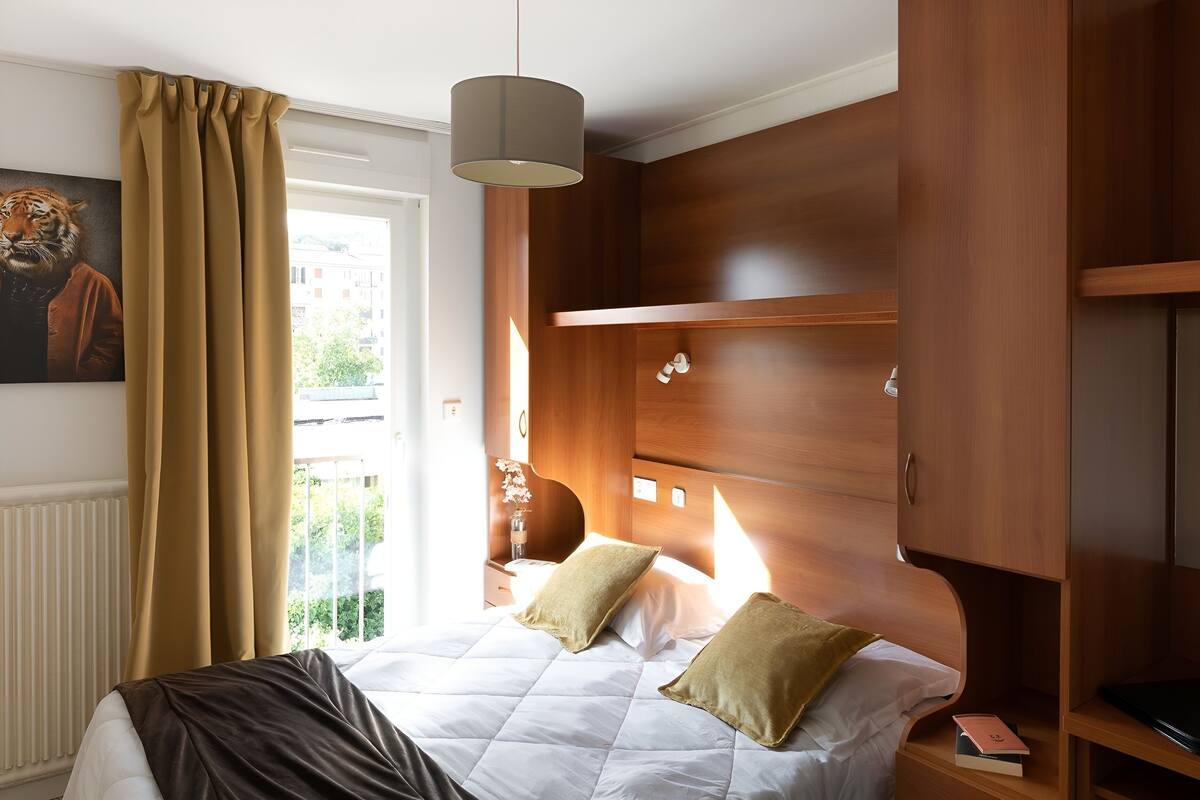 Un dormitorio con cama, mesita de noche, armario de madera, ventana con cortinas y un cuadro en la pared.