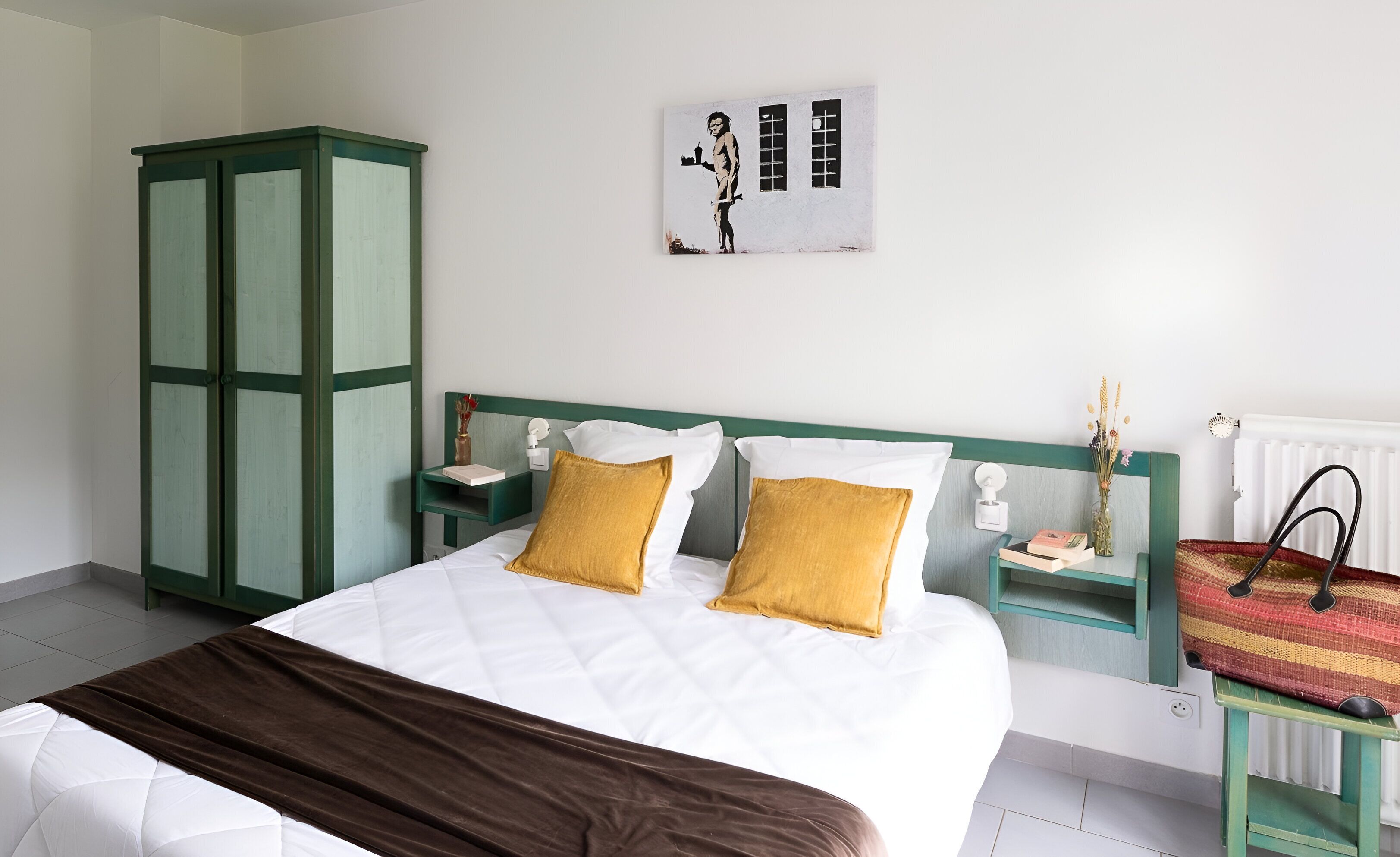 Un dormitorio con una cama, un armario verde, mesitas de noche, una obra de arte colgada en la pared y un radiador.