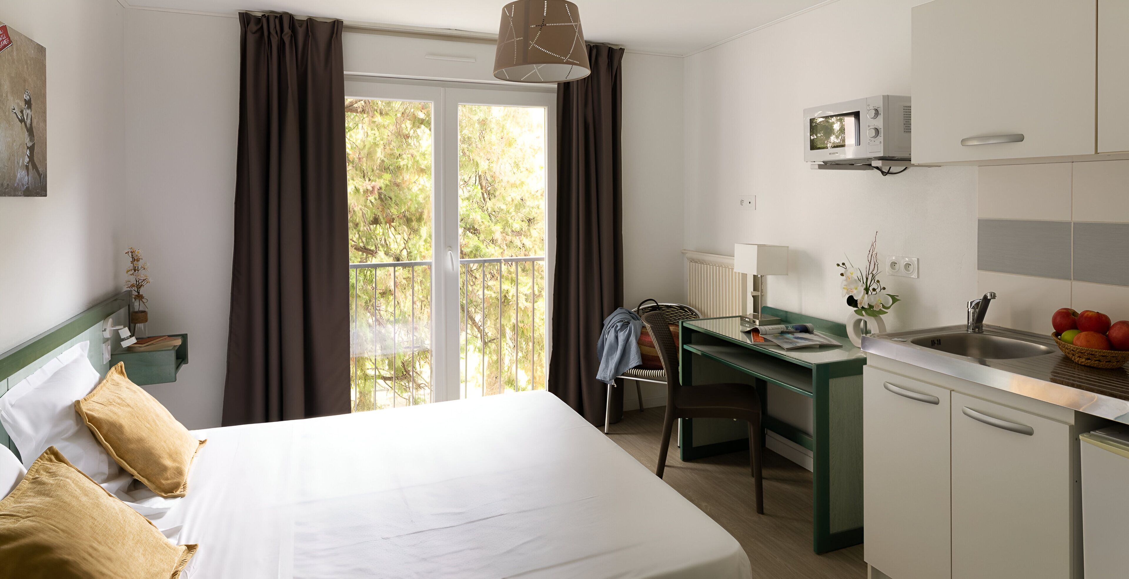 Ein Hotelzimmer mit Bett, Schreibtisch, Stuhl, einer Küchenzeile und Blick auf Bäume durch das Fenster.