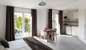 A hotel room with a bed, a dining table with breakfast, a kitchenette, and large windows with curtains.