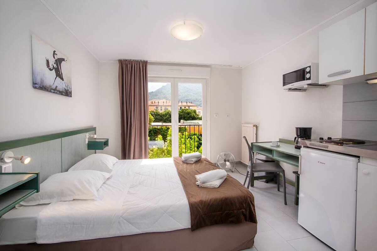 Habitación de hotel con cama, escritorio, kitchenette y vistas a edificios y zonas verdes.