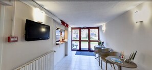 Un pasillo con un televisor montado en la pared, una recepción y una zona de estar con sillas y una mesa.