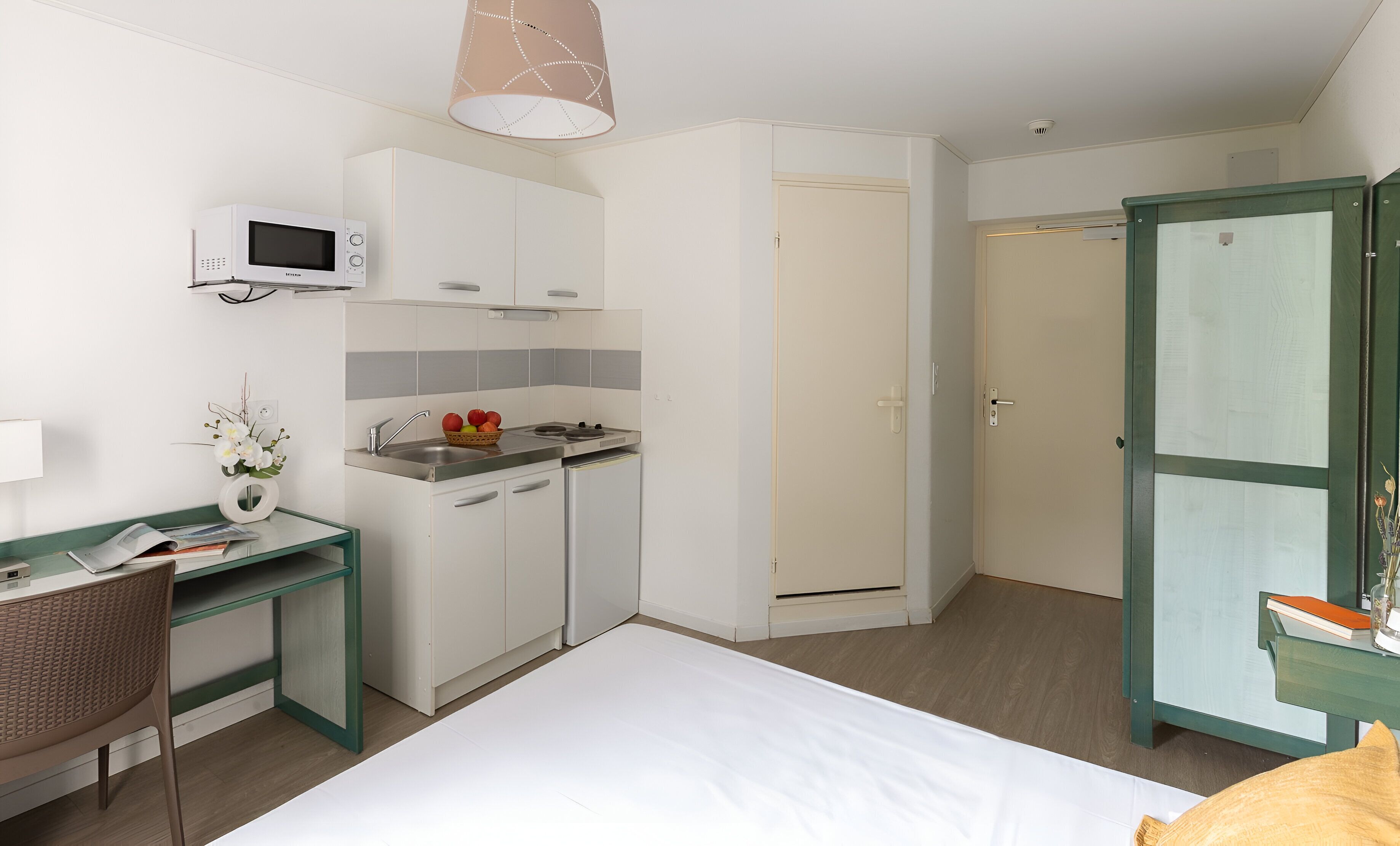 Ett kompakt hotellrum med ett litet kök, en säng, ett skrivbord med en stol och en garderob.