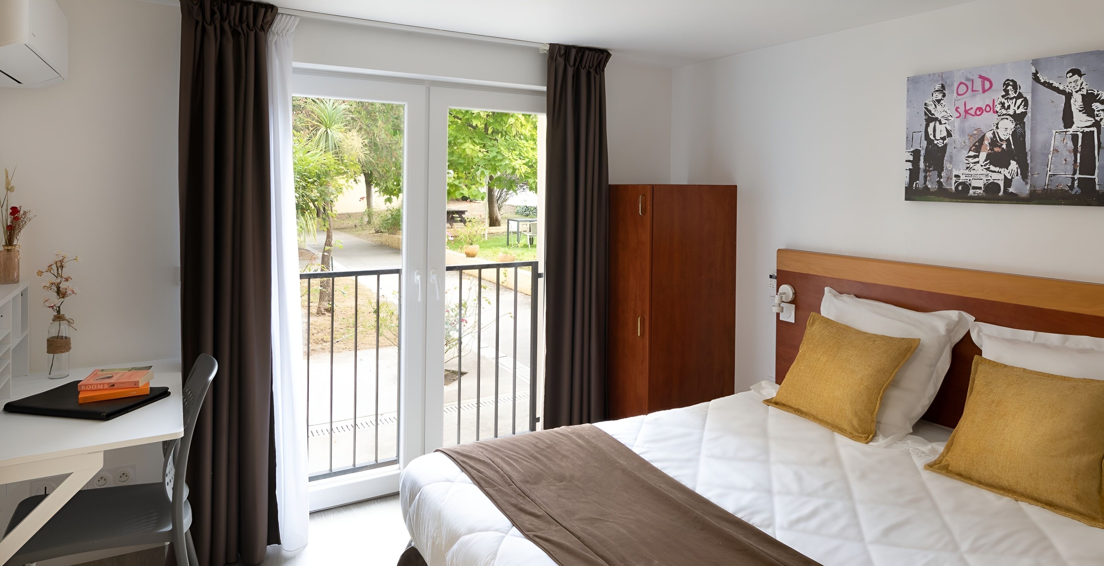 A hotel room with a bed, a desk, a chair, a wardrobe, and a balcony with a view of greenery.