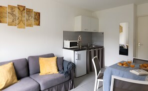 A modern kitchenette with a microwave, sink, and cabinets. There is a gray sofa with a yellow pillow and a blue blanket. A dining table with a blue tablecloth and a basket of croissants is visible. - Appart Hôtel Corte HR (Corte)