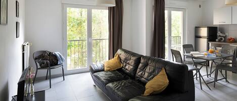 A modern living room with a leather sofa, dining table, chairs, and a kitchen area with a refrigerator.