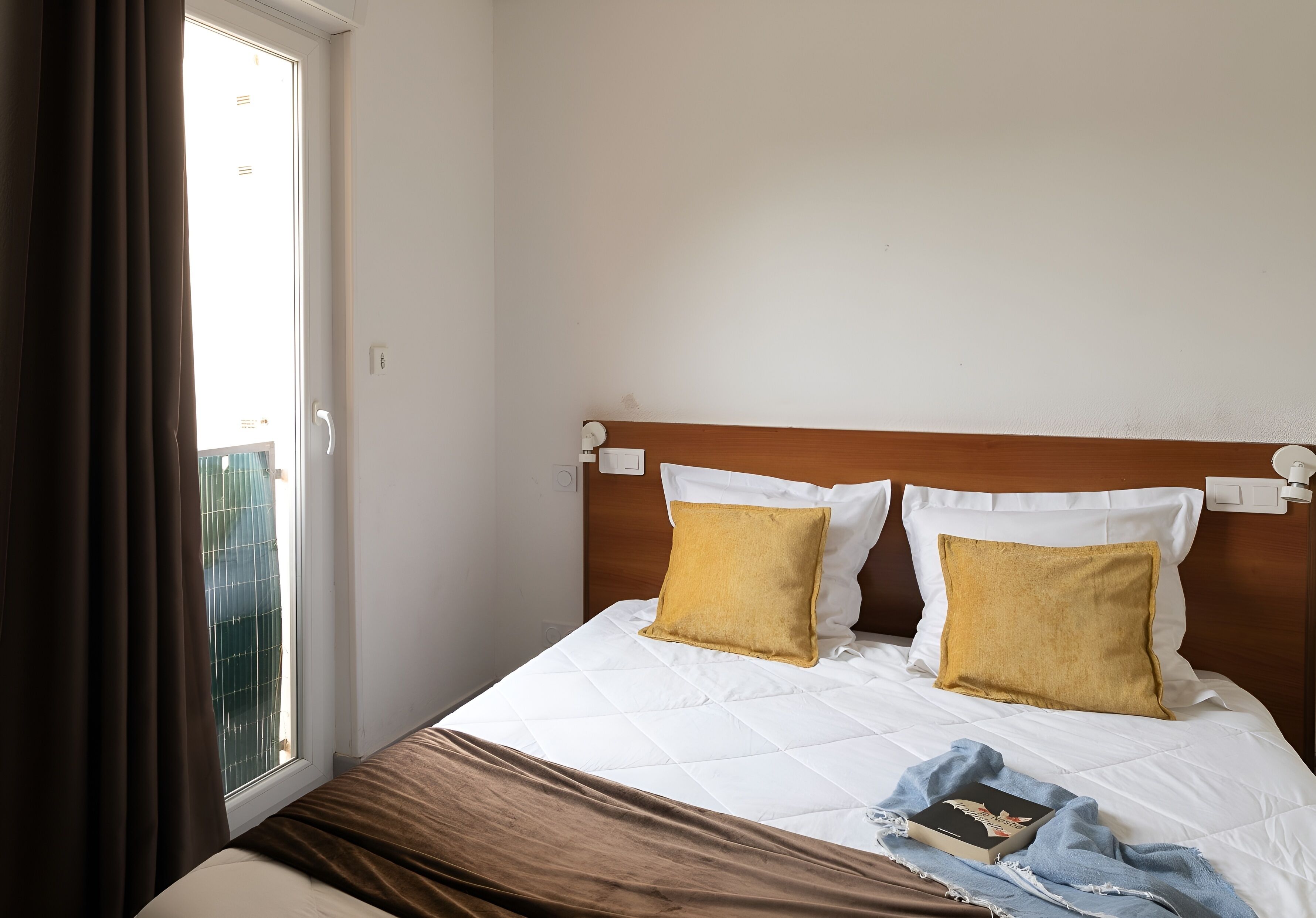 A hotel room with a bed, two pillows, a blanket, and a book.