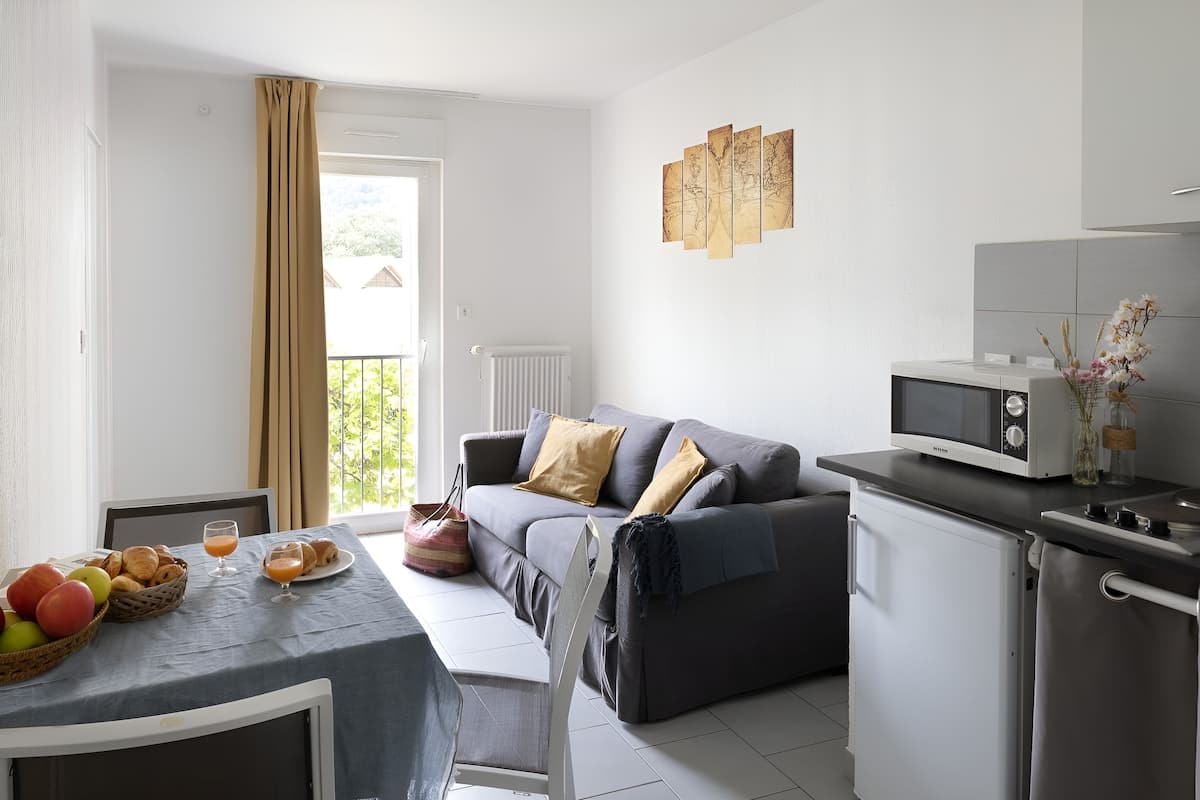 Una cocina moderna con un sofá gris, una mesa de comedor con frutas y croissants, un refrigerador pequeño y un microondas.