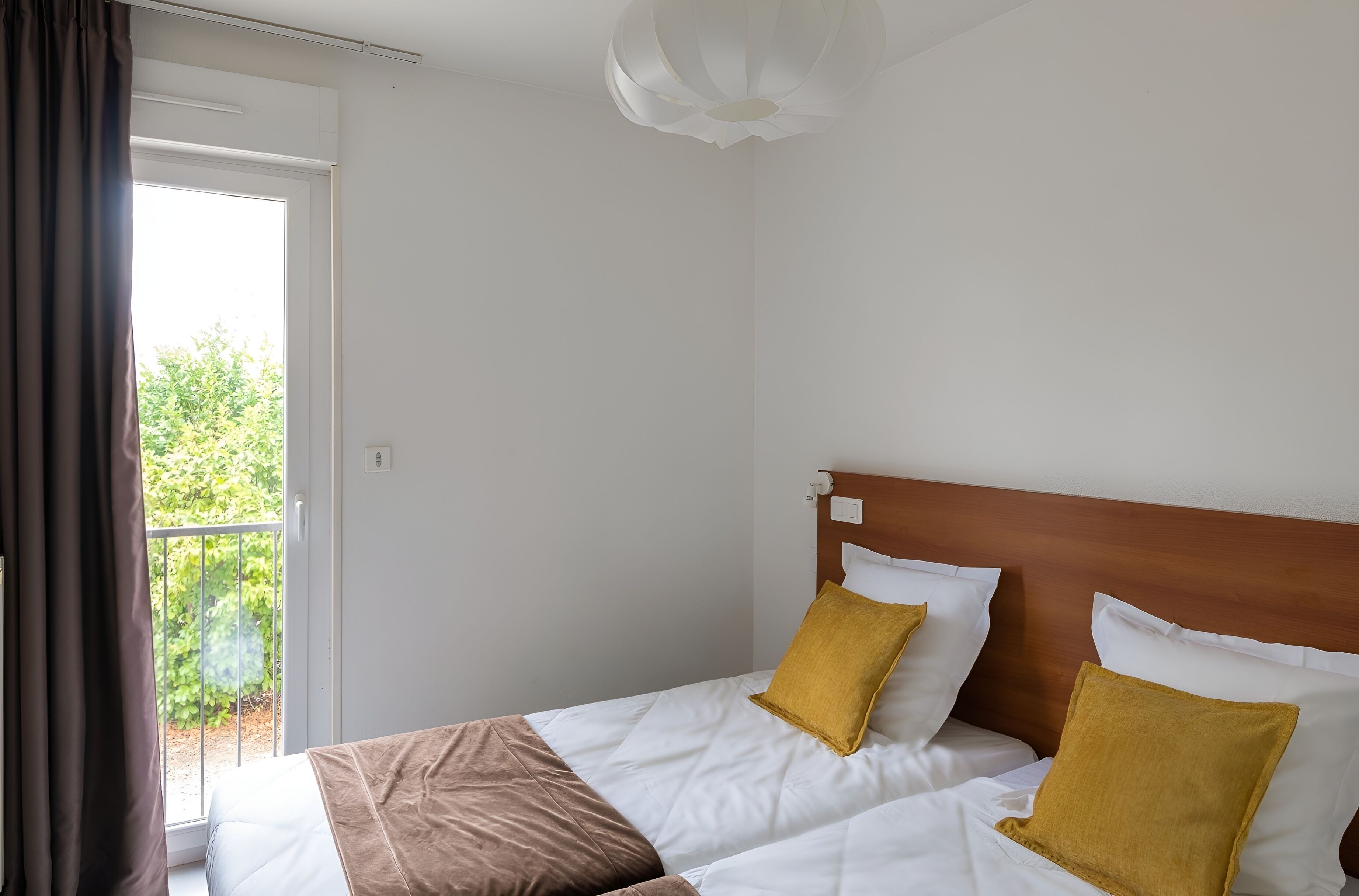 A hotel room with a bed, a wooden headboard, and a view of greenery outside.