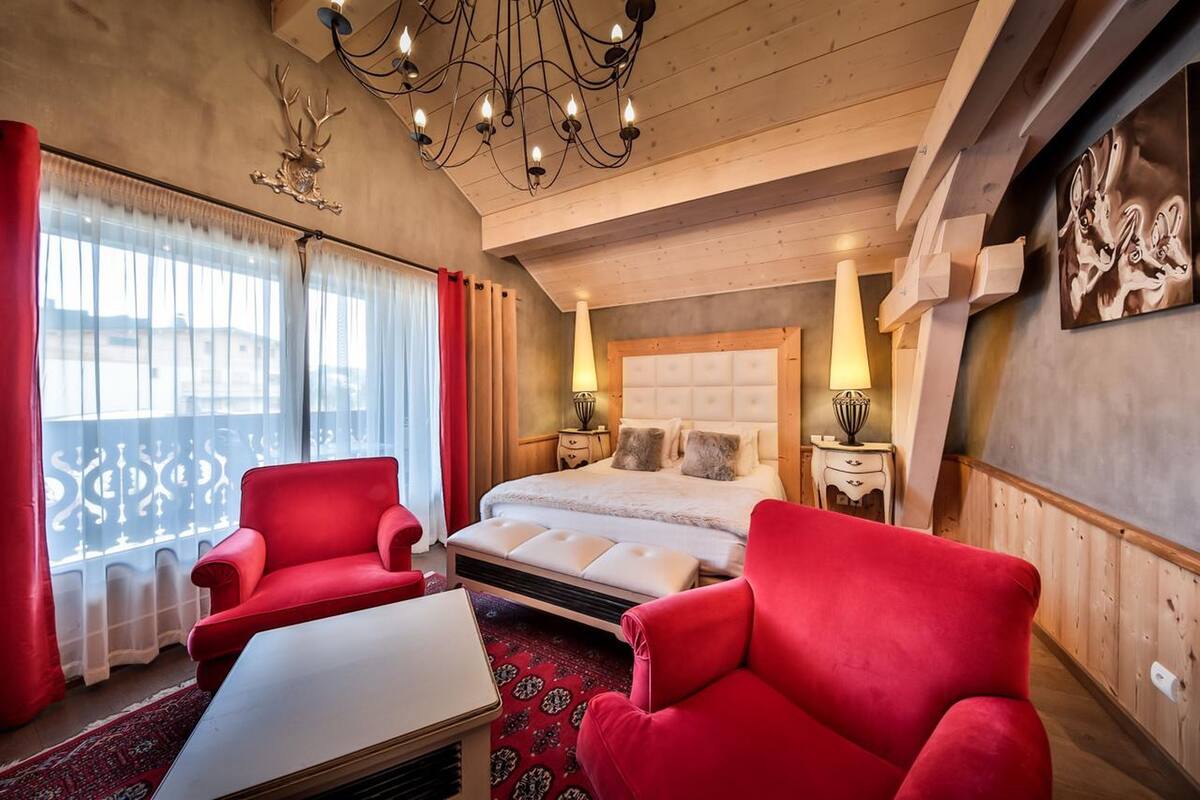 Ein Schlafzimmer mit einem großen Bett, zwei roten Sesseln, einer Chandelieber und Blick ins Freie durch das Fenster.
