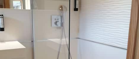 Un espace douche clos par des parois de verre, équipé d’un pommeau de douche à main, d’un panneau de commande et d’un petit drain.