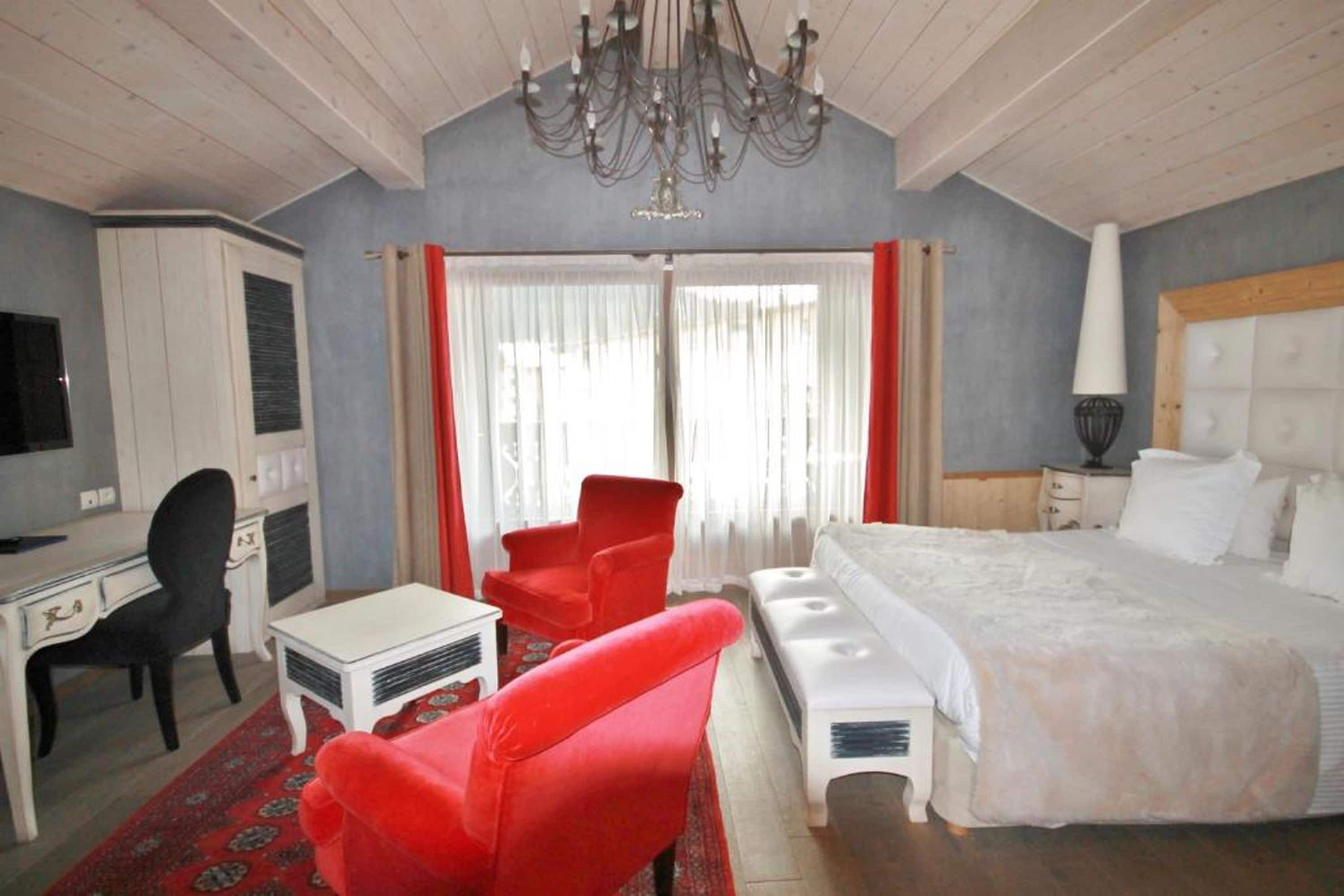 A bedroom with a bed, a desk, a chair, a red armchair, and a chandelier.