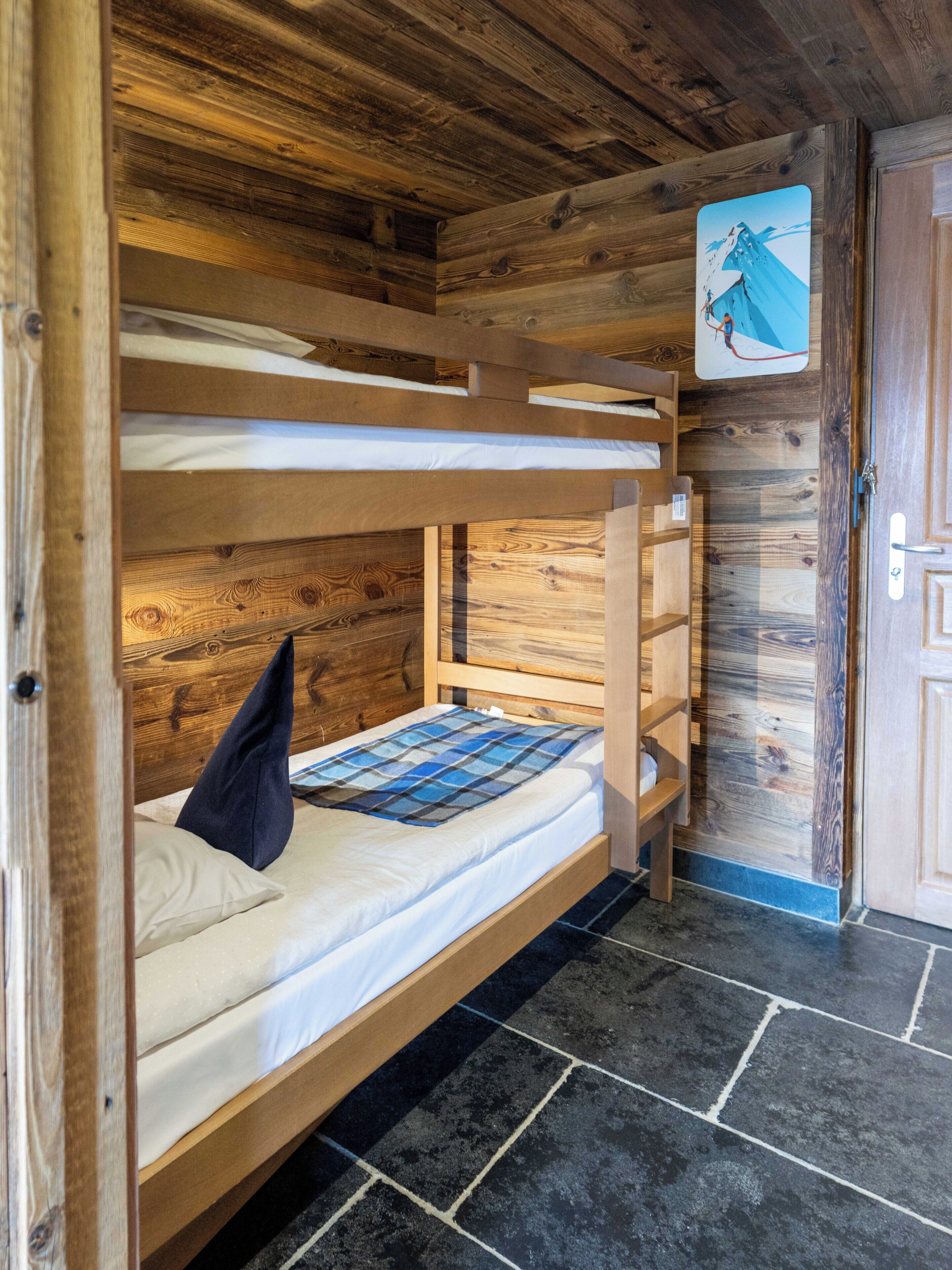 A bunk bed with a blue and white checkered blanket, wooden walls, and a door with a mountain painting.