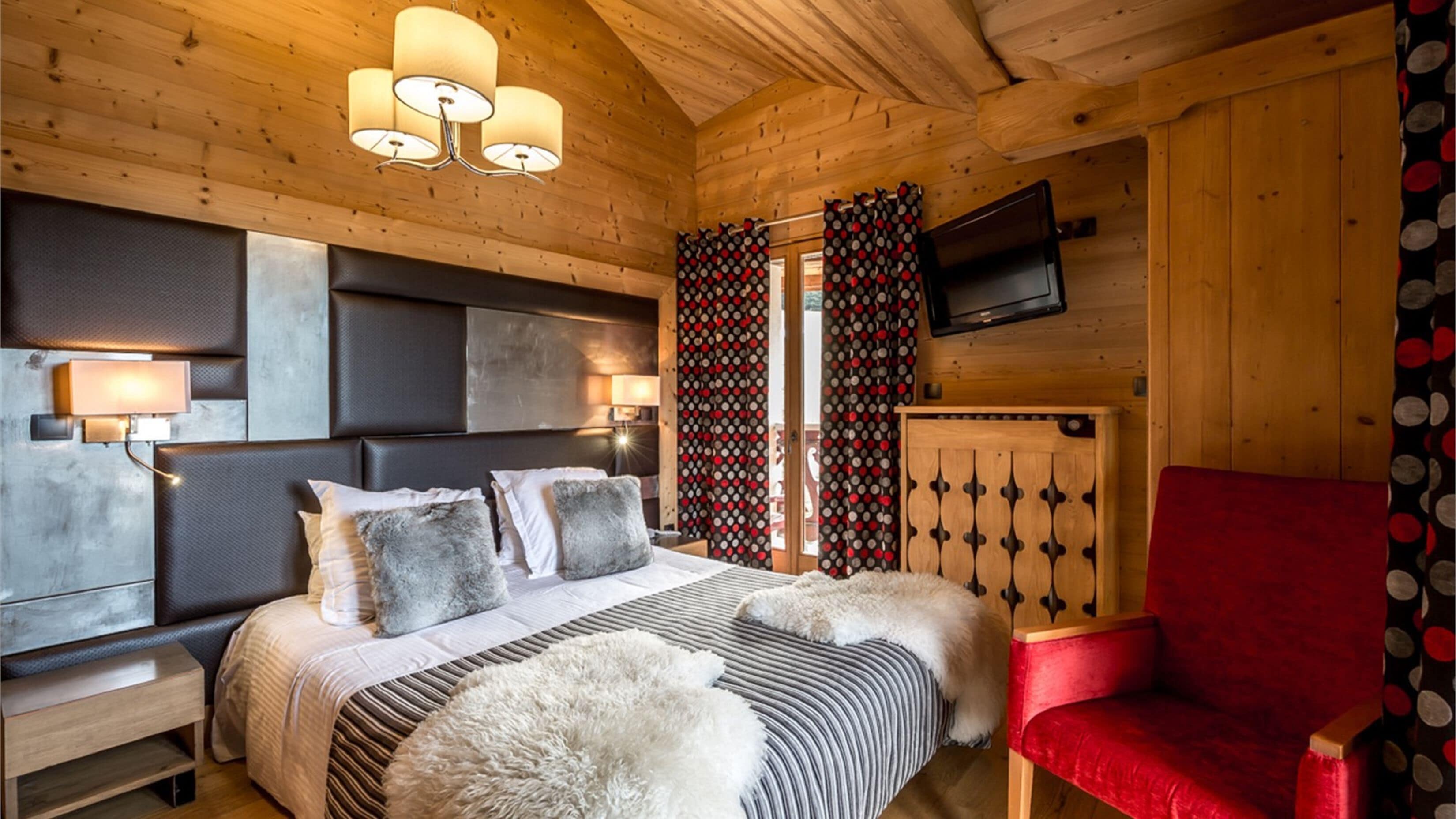 A wooden cabin bedroom with a bed, a red chair, a TV, and a patterned curtain.