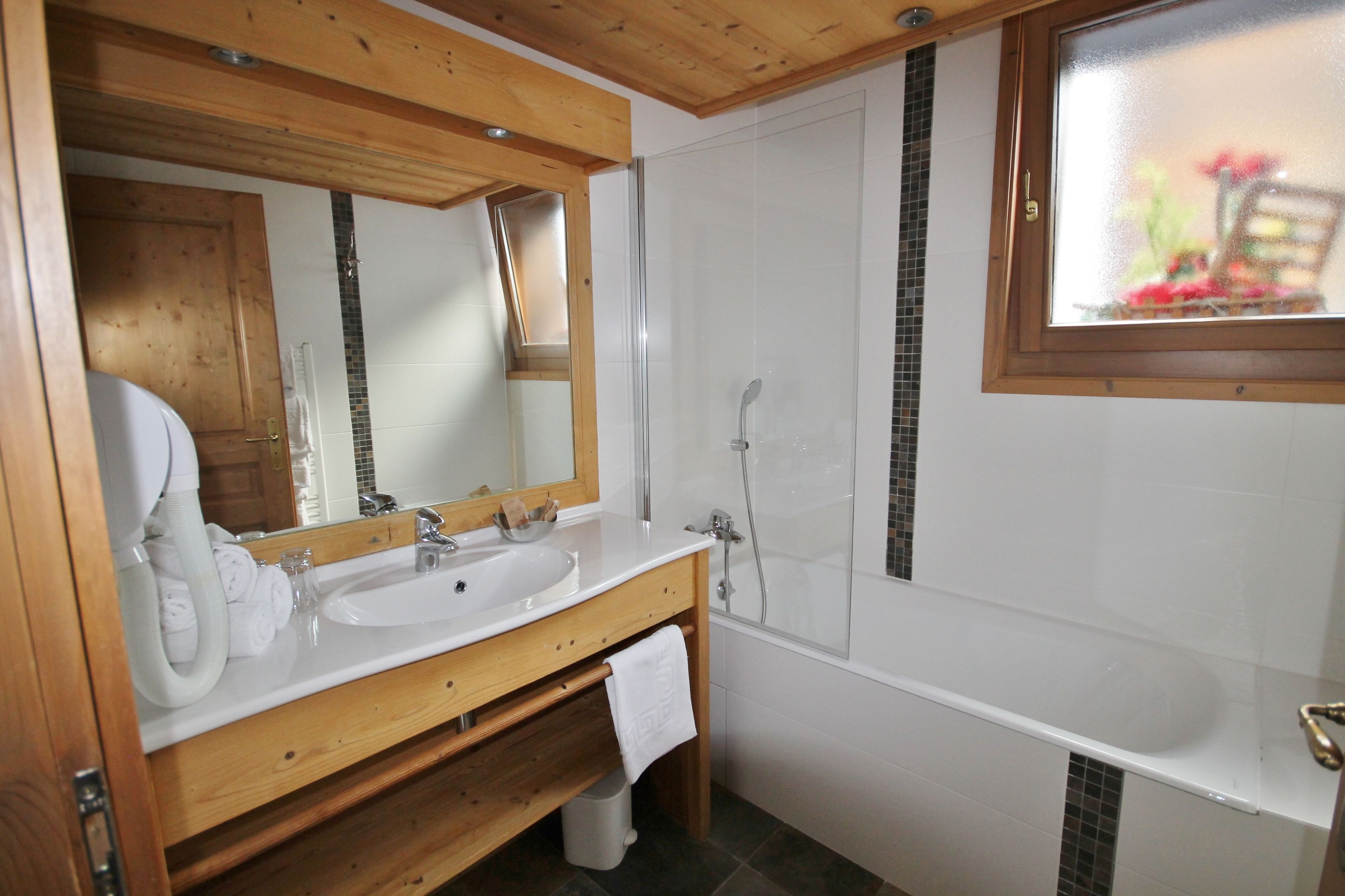 A bathroom with a wooden vanity, a large mirror, a white sink, a bathtub, and a window with a view of flowers.