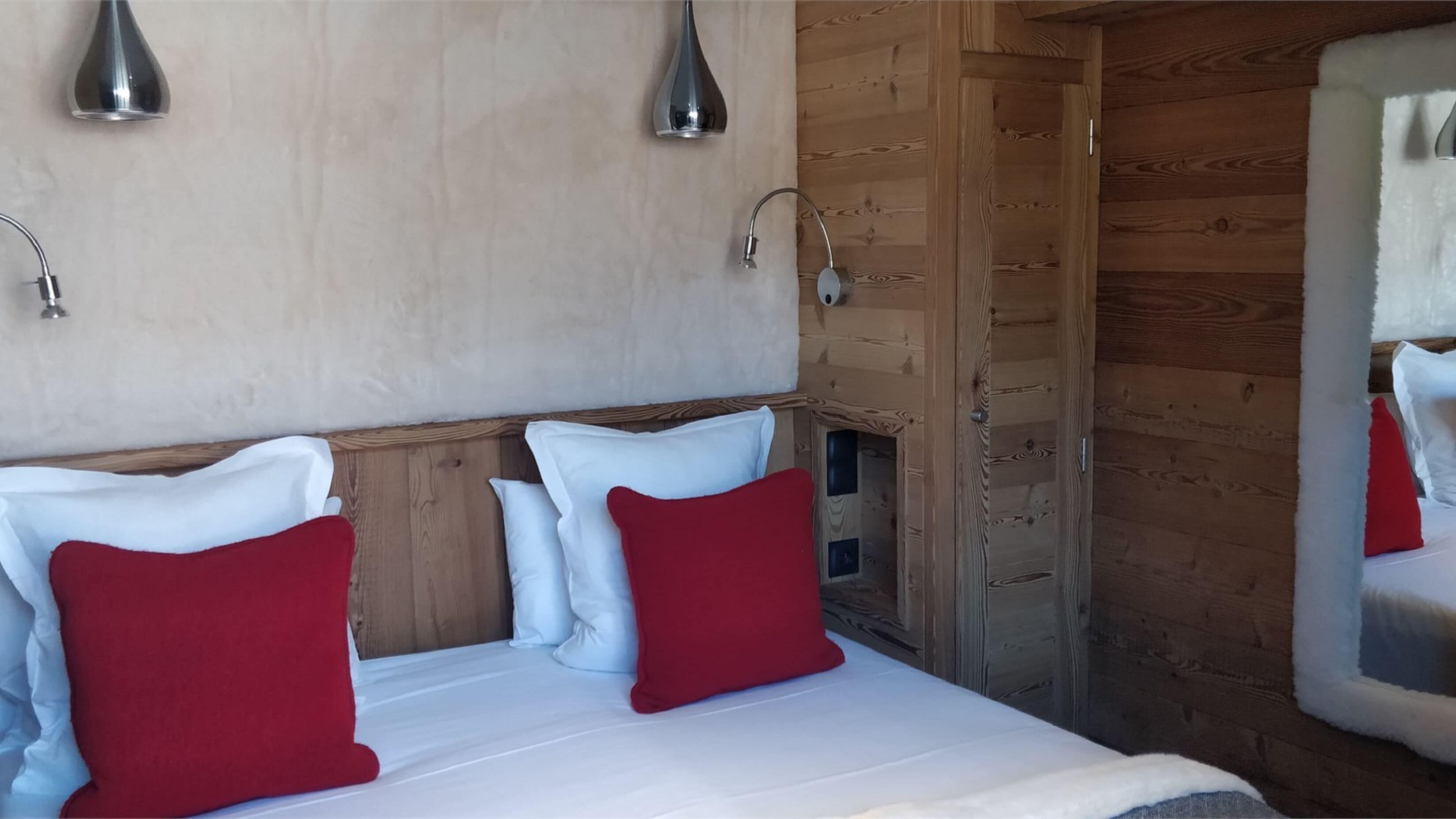 Interior de una cabaña de madera con una cama, dos almohadas rojas y dos lámparas fijadas a la pared.