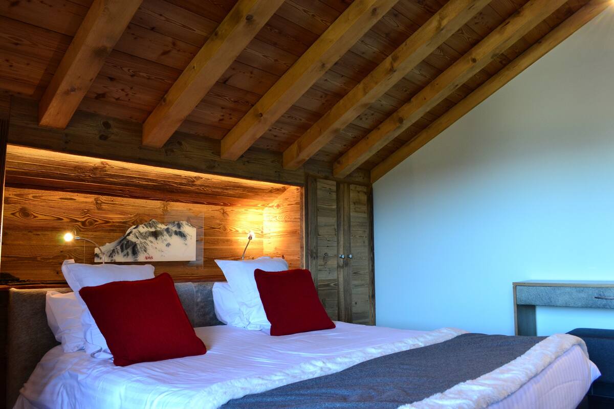 Eine Holzbalkendecke, ein Bett mit weißer Bettwäsche und roten Kissen sowie eine Wandleuchte.