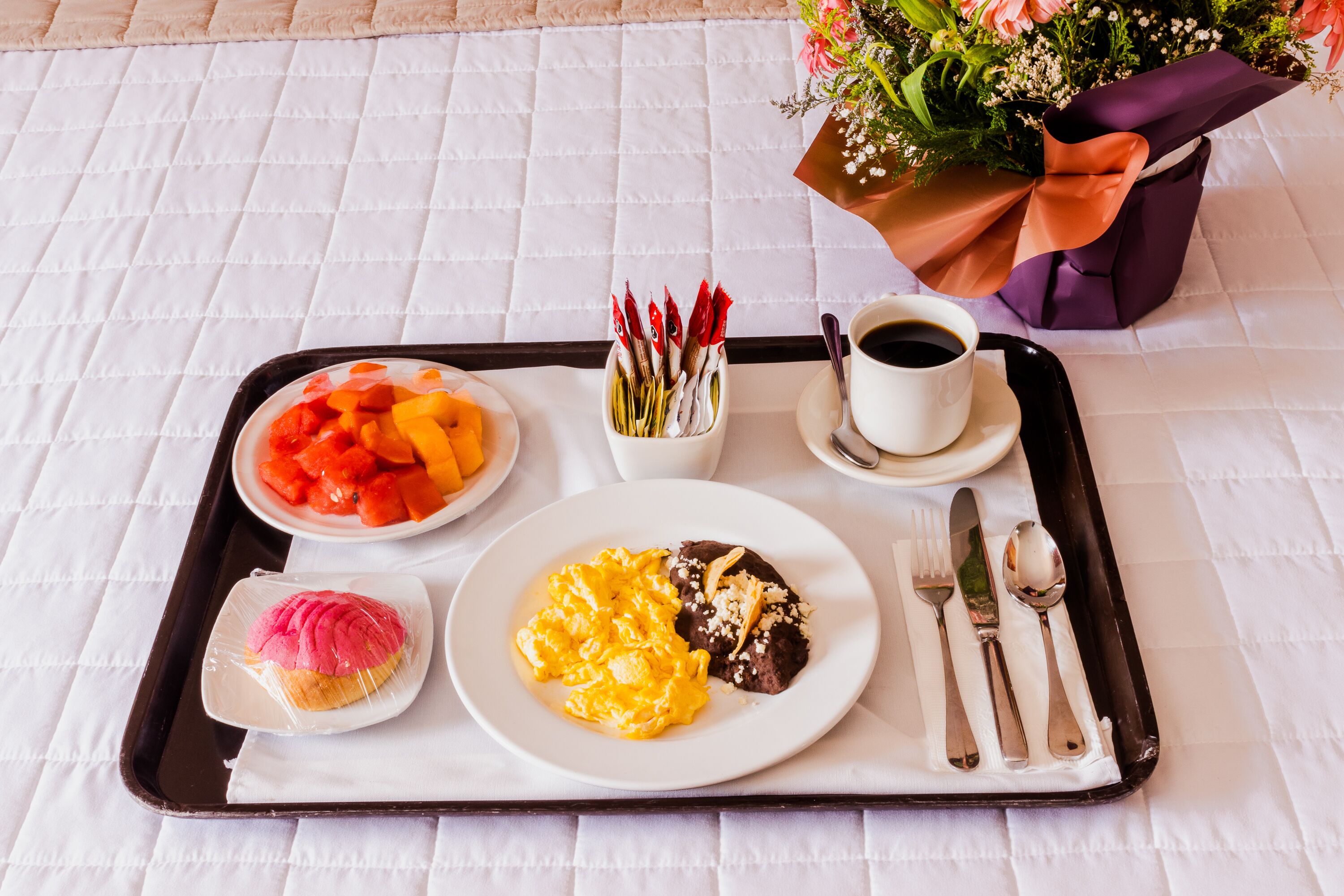 Bandeja con alimentos para desayuno, incluyendo fruta, huevos revueltos y un postre, además de una taza de café y cubiertos sobre una cama con colcha blanca acolchada.