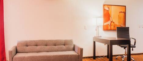 A room with a sofa, a desk, a chair, an air conditioner, and a painting on the wall.