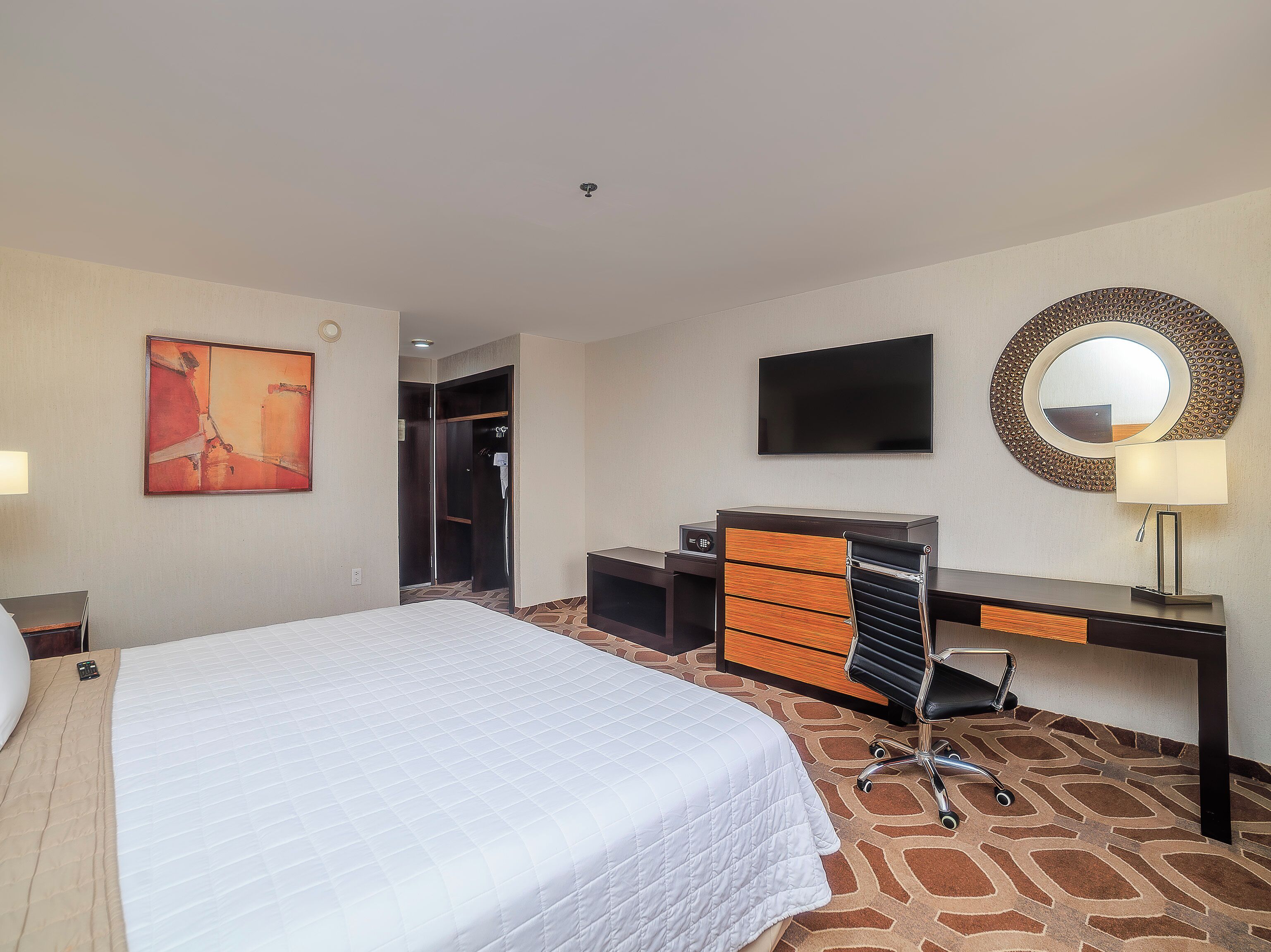 A hotel room with a bed, a desk with drawers, a television, a mirror, and a painting on the wall.