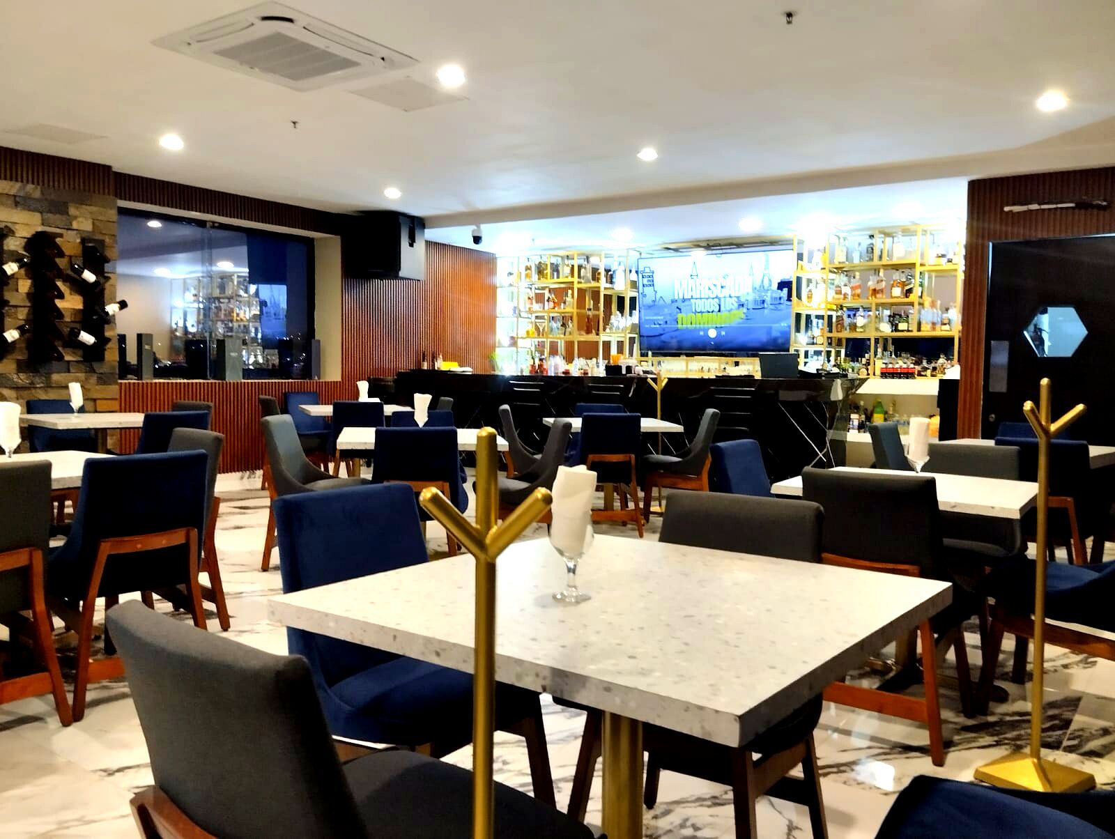 A restaurant with marble-topped tables and chairs, a bar area with shelves of bottles, and a television screen displaying a game.