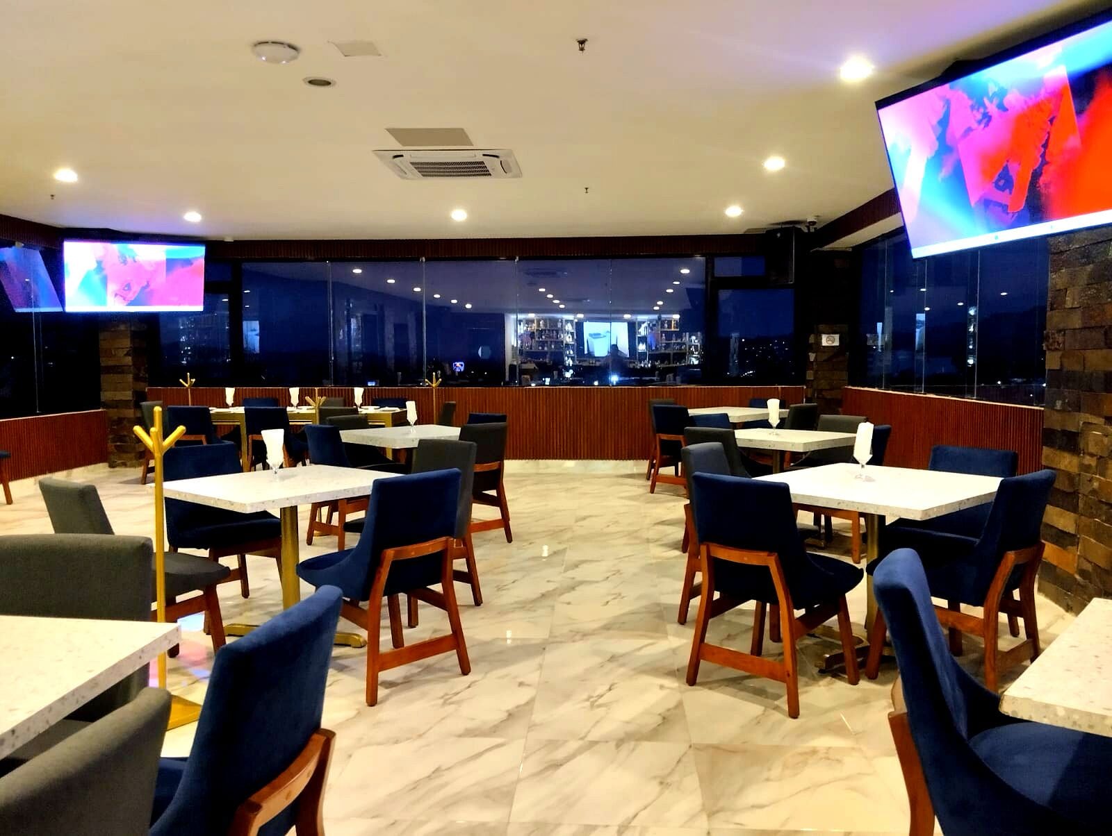Un restaurante con varias mesas y sillas, una zona de bar y grandes ventanales que ofrecen vistas del paisaje urbano.
