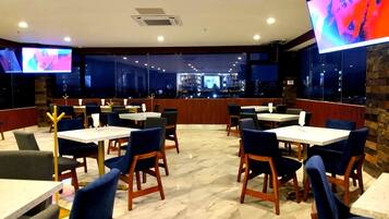 Un restaurante con varias mesas y sillas, una zona de bar y grandes ventanales que ofrecen vistas del paisaje urbano.